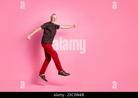 Foto a grandezza naturale di una t-shirt marrone da uomo senior con abiti positivi, pantaloni rossi che stringono il pugno a volo vuoto, isolata su sfondo di colore rosa Foto Stock