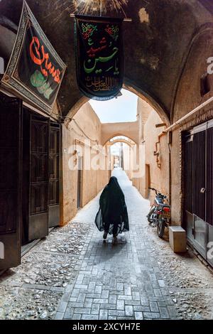 Yazd, Iran - 26 ottobre 2018: Donna iraniana che indossa un chador nero che cammina lungo le strette strade della storica città di Yazd. Foto Stock