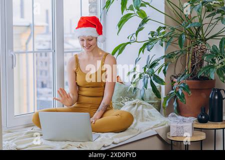 Donna che indossa un cappello di Babbo Natale e un abito color senape, siede accanto alla finestra e videochiama sul suo laptop. È circondata da piante al coperto e decorazioni natalizie, creando un'atmosfera festosa e accogliente. Foto Stock