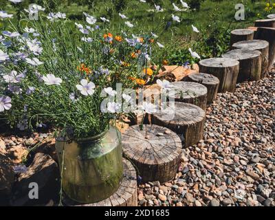 Un vaso di vetro pieno di fiori bianchi poggia su un tronco di legno in un ambiente naturale. Il percorso del tronco conduce attraverso un letto di ghiaia con altri fiori in fiore sullo sfondo. Foto Stock