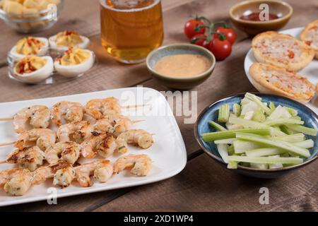 tavolo con spiedini di gamberi grigliati, uova diavolate, bastoncini di sedano e un bicchiere di birra. Perfetto per incontri sociali, bar e feste informali Foto Stock