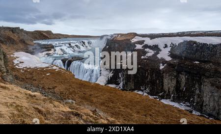 Una vista mozzafiato della cascata Gullfoss in Islanda, circondata da scogliere innevate e terreno accidentato sotto un cielo nuvoloso. Foto Stock