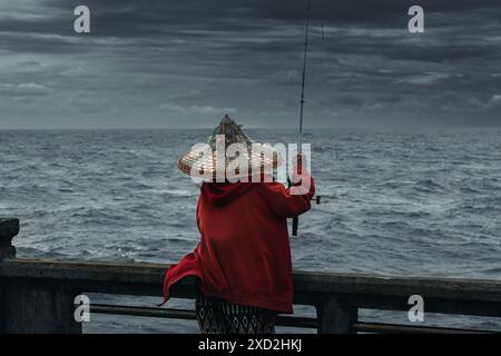 Donne con canna da pesca che pescano pesce in Thailandia. Donna asiatica con giacca rossa che pesca sul porto durante la tempesta. pesca durante la stagione delle piogge Foto Stock