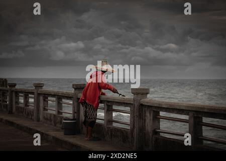 Donne con canna da pesca che pescano pesce in Thailandia. Donna asiatica con giacca rossa che pesca sul porto durante la tempesta. pesca durante la stagione delle piogge Foto Stock