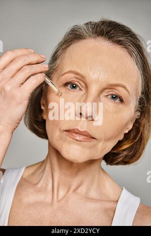 Una donna matura e attraente in un abbigliamento comodo le applica sapientemente il siero sul viso. Foto Stock