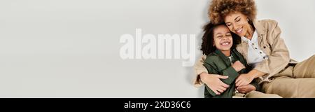 Una madre riccia afro-americana abbraccia amorevolmente sua figlia, entrambe in abiti eleganti, su uno sfondo grigio. Foto Stock
