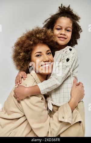 Madre e figlia ricci afroamericani in abiti eleganti che si abbracciano amorevolmente su uno sfondo grigio. Foto Stock