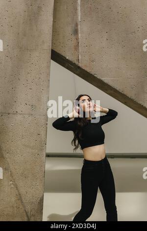 Una giovane donna sta ascoltando la musica attraverso le sue cuffie, appoggiandosi alla schiena contro una parete di cemento testurizzato in un ambiente urbano Foto Stock