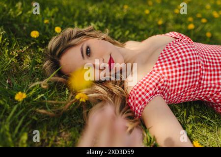 La giovane donna con un abito a scacchi rosso gode di un momento di tranquillità in un campo verde punteggiato di fiori gialli Foto Stock