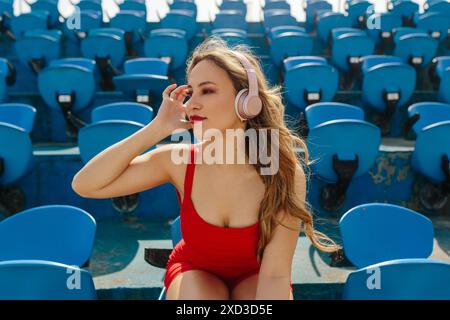 Una giovane donna con un costume da bagno rosso siede sui sedili blu dello stadio e ascolta la musica con le cuffie bianche in una giornata di sole Foto Stock