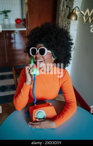 Una gioiosa donna afro-americana nera vestita con dolcevita arancione ispirata agli anni '1950 e accessoriata con grandi occhiali da sole bianchi e un cerchietto, Laugh Foto Stock