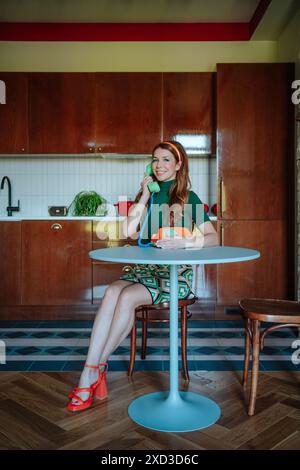 Una donna rossa sorridente vestita in stile anni '50 siede a un tavolo da cucina, chattando comicamente su un classico telefono verde, evocando un nostalgico su Foto Stock