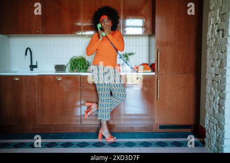 Una donna afroamericana nera vestita con un vivace abbigliamento anni '50 appare scioccata mentre parla su un classico telefono verde in un ambiente da cucina vintage. Foto Stock