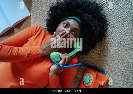 Dall'alto, la giovane donna afroamericana nera vestita con un maglione arancione brillante giace sul pavimento, guardando la macchina fotografica con un espresso sorpreso Foto Stock