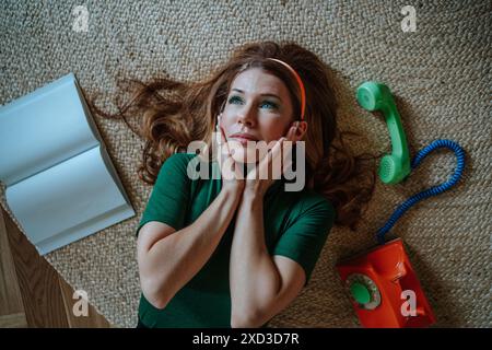 Dall'alto, donna premurosa in una camicia verde, sdraiata su un tappeto che sfoggia un'espressione riflessiva accanto a una macchina da scrivere retrò arancione e a un verde Foto Stock