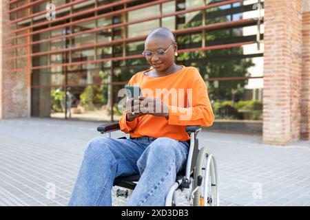 Donna afroamericana sicura in sedia a rotelle, con un maglione arancione, che si impegna con uno smartphone in un ambiente urbano Foto Stock