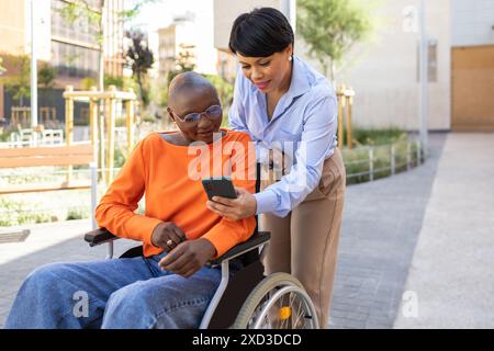 Una donna d'affari aiuta il suo collega afro-americano, che è su una sedia a rotelle, a utilizzare uno smartphone all'aperto, entrambi sono impegnati e sorridenti Foto Stock