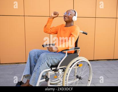 Una donna afroamericana in sedia a rotelle ascolta la musica con le cuffie, sentendosi gioiosa e sollevata, all'aperto con un moderno sfondo arancione Foto Stock