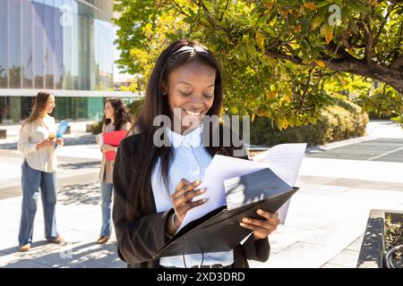 Una donna d'affari di colore è impegnata a rivedere i documenti fuori da un ufficio urbano, con due colleghi di sesso femminile che discutono sullo sfondo sotto Foto Stock