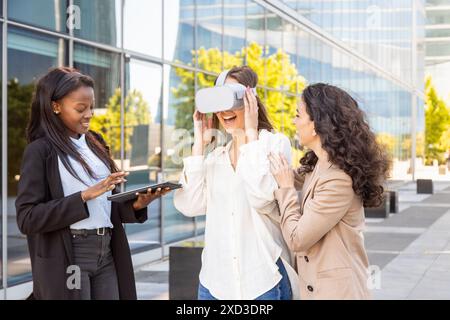 Tre donne professionali multirazziali si impegnano con la tecnologia della realtà virtuale per una dimostrazione aziendale all'esterno dei moderni edifici adibiti a uffici Foto Stock