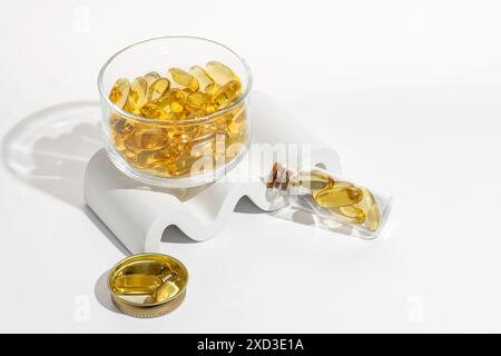 Capsule Omega 3 presentate elegantemente in una ciotola di vetro trasparente con alcune capsule versate su un piedistallo bianco ideale per temi di salute e benessere Foto Stock