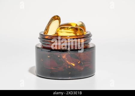 Primo piano di capsule dorate di olio di pesce Omega 3 che fuoriescono da un vaso di vetro color ambra scuro su una superficie bianca liscia Foto Stock