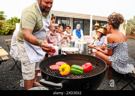 Gruppi diversi di amici che amano la musica e un barbecue all'aperto. Un uomo grigia carne e verdure mentre i partecipanti multietnici si impegnano in una conversazione. Tutti Foto Stock