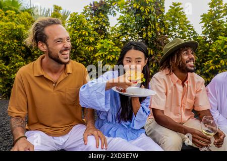 Un gruppo eterogeneo di amici condivide risate e cibo a un barbecue all'aperto. Una donna asiatica mangia un hamburger, un uomo caucasico ride, e un Ameri africano Foto Stock