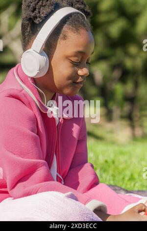 Una giovane ragazza nera che indossa una giacca rosa e le cuffie ama ascoltare la musica mentre è seduta su una coperta bianca in un parco illuminato dal sole Foto Stock