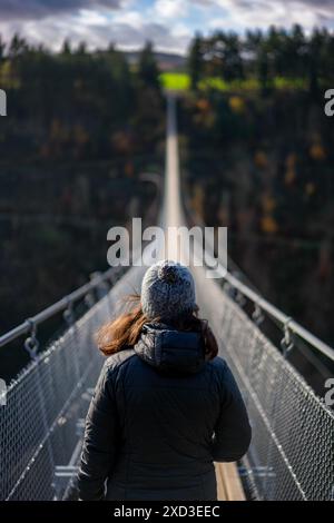 Vista posteriore di una donna irriconoscibile che cammina attraverso il ponte sospeso di Geierlay, uno dei ponti di corda più lunghi della Germania, circondato da foreste autunnali Foto Stock