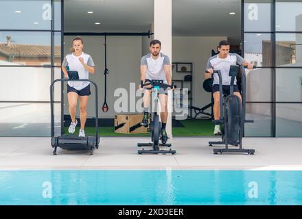 Tre giovani adulti sono impegnati in una sessione di allenamento in una moderna palestra all'aperto che si affaccia su una piscina e utilizzano varie attrezzature per il fitness come il treadmil Foto Stock