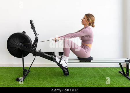 Una giovane donna concentrata nell'abbigliamento da palestra rosa coordinato si sta allenando su una moderna vogatrice in uno spazio palestra illuminato con erba verde artificiale Foto Stock