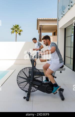 Due uomini si sono concentrati sull'allenamento utilizzando una cyclette e un tapis roulant in una palestra all'aperto con vista panoramica sulle palme Foto Stock