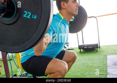 Un uomo concentrato di abbigliamento sportivo blu e giallo esegue uno squat, tenendo in mano un barbell carico di pesi da 22 kg in un ambiente luminoso e moderno Foto Stock