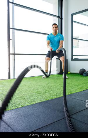 Un atleta esegue un intenso allenamento con corda da battaglia in una palestra con interni luminosi e moderni, con ampie finestre e un pavimento verde verdeggiante Foto Stock