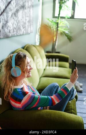 Una giovane donna allegra si gode una pausa su un divano verde, ascoltando musica con le cuffie e utilizzando lo smartphone in una stanza piena di luce Foto Stock