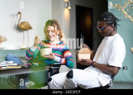 Una giovane coppia felice e multietnica si prende una pausa dal lavoro intelligente a casa, gustando un pasto insieme in un ambiente accogliente e moderno con illustrazioni della natura Foto Stock