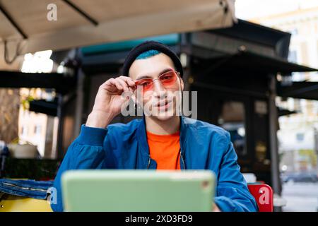 Un giovane uomo alla moda con una giacca blu e una camicia arancione tocca i suoi occhiali da sole color rosa mentre è seduto con un computer portatile in un bar all'aperto Foto Stock