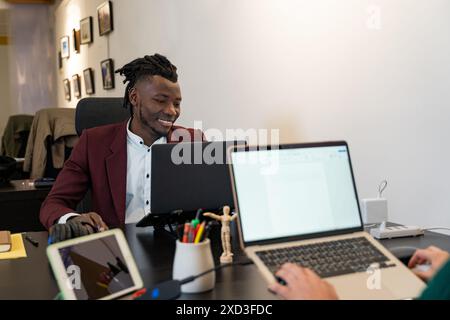 Un giovane uomo d'affari allegro in un elegante blazer color marrone lavora con attenzione sul suo notebook, interagendo con un collega di un ufficio moderno e ben attrezzato Foto Stock
