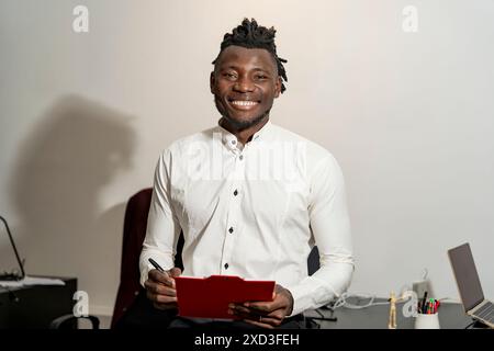 Un giovane professionista, sorridente con fiducia, si trova davanti a una scrivania mentre tiene in mano gli appunti, indossa una camicia bianca nitida, che segnala un lavoro informale Foto Stock