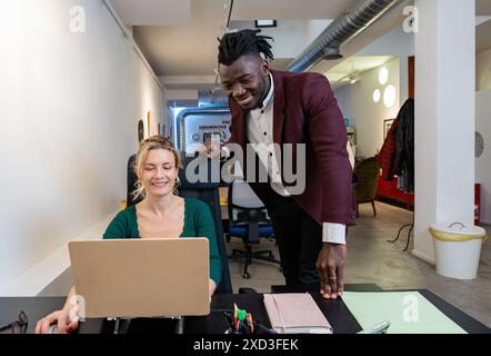 Una giovane donna si siede a una scrivania, sorridendo mentre guarda un notebook accanto a lei, un giovane uomo con un blazer color marrone e una camicia nera punta sullo schermo, givin Foto Stock