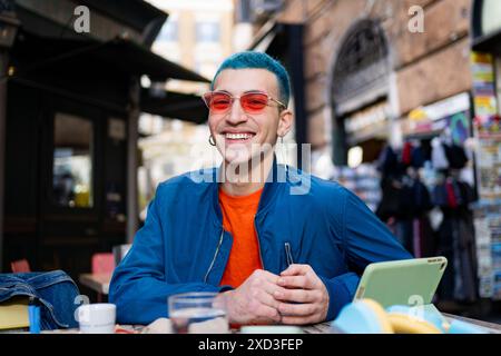 Giovane uomo felice con capelli blu vivaci e eleganti occhiali da sole rossi si siede al tavolo di un bar, trasudando gioia mentre si impegna con il suo smartphone, surround Foto Stock