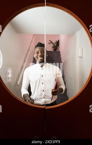 Un allegro uomo nero in una camicia bianca, riflessa in uno specchio circolare, tiene i documenti mentre si trova in piedi su scale con pareti rosa Foto Stock