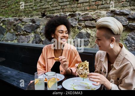 Due donne diverse e belle si siedono a un tavolo e mangiano felicemente pizza in un accogliente bar. Foto Stock