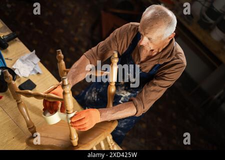 Un uomo anziano con guanti arancioni e grembiule ripara una vecchia sedia di legno in un'officina. Avvolge le gambe della sedia con del nastro prima di dipingere. Foto Stock