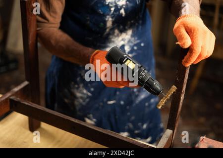 mani con guanti arancioni e grembiule blu che riparano una sedia con un trapano elettrico in un'officina. Levigare la sedia con un accessorio e rimuovere un Foto Stock