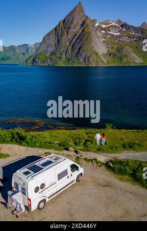 Un camper bianco parcheggiato vicino a una coppia godendo della vista di uno splendido fiordo norvegese e della catena montuosa. L'acqua blu e le verdi colline creano una scena pittoresca. Lofoten Norvegia Foto Stock