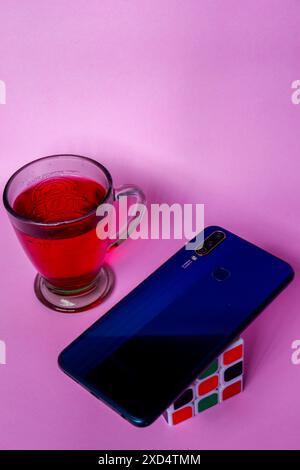 Foto di bevanda rossa, smartphone blu e Rubik su sfondo rosa Foto Stock