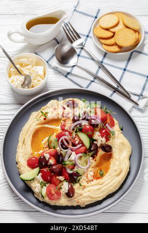 Hummus greco carico di cetrioli freschi, olive, pomodori, feta, cipolle rosse e timo su un tavolo di legno bianco con cracker, V verticale Foto Stock