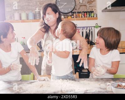 I figli gettano la farina in faccia alla madre mentre preparano la ricetta della torta in cucina. Foto Stock